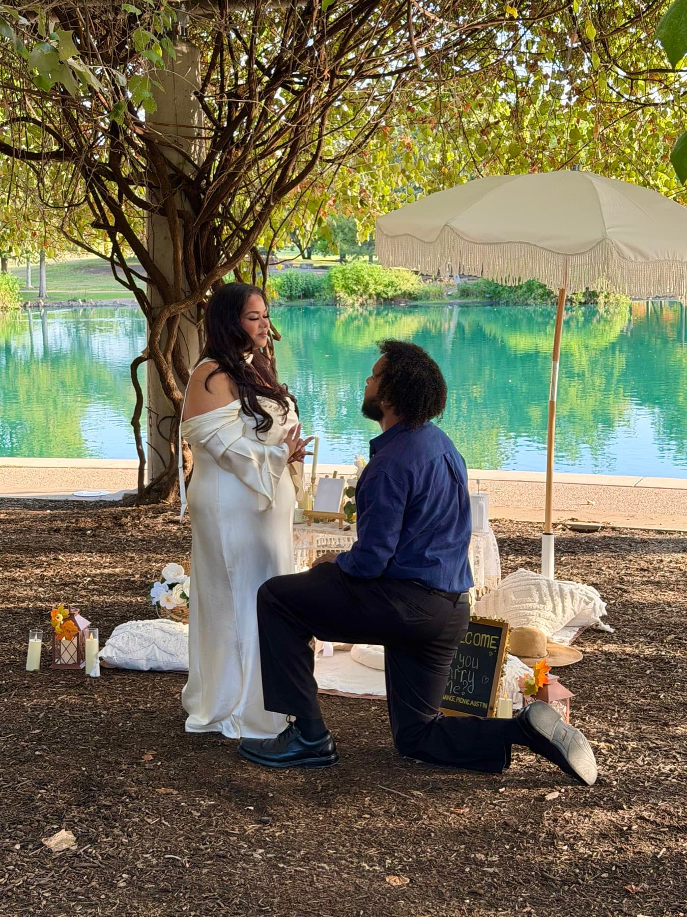 Man on one knee proposing at a lakeside picnic with a fringe umbrella in Austin